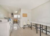 Thumbnail 9 of 14 - A laundry room with a white washer and dryer, a white chair, and a white table.