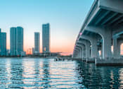 Thumbnail 67 of 71 - A city skyline is reflected in the water under a bridge.