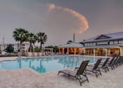 Thumbnail 6 of 26 - A pool surrounded by lounge chairs with a building and palm trees in the background.