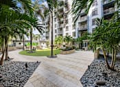 Thumbnail 30 of 63 - A courtyard with a paved walkway and a grassy area surrounded by palm trees and apartment buildings.