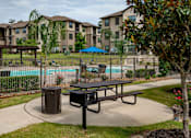 Thumbnail 13 of 28 - A park area with a picnic table and a trash can in front of apartment buildings.