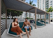 Thumbnail 29 of 76 - A man and a woman are sitting on a bench under a shaded area.