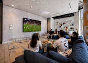 Thumbnail 17 of 76 - A group of people are watching a soccer game on a large TV screen in a modern living room.