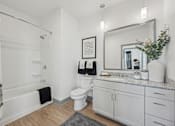 Thumbnail 14 of 26 - A bathroom with a white toilet, a shower with a curtain, a mirror, and a plant on the counter.