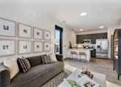 Thumbnail 11 of 27 - A modern living room with a grey couch and a coffee table in front of a wall with framed pictures.