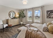 Thumbnail 14 of 45 - Sunlit primary bedroom with large double windows, plaid drapes, natural wood dresser with round gold mirror, olive tree accent, white upholstered accent chair with leather footstool, botanical wall art, and cozy layered bedding in warm neutral tones