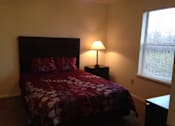 Thumbnail 28 of 28 - A bed with a red and white comforter sits in a room with a lamp on a side table.
