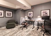 Thumbnail 9 of 20 - A room with grey walls and a carpeted floor with a green armchair, two desks with chairs, and framed pictures on the wall.