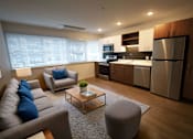 Thumbnail 32 of 33 - A modern living room with a grey couch, a coffee table, and a kitchen area in the background.