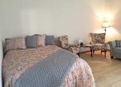 Thumbnail 4 of 10 - A bed with a grey comforter and matching pillows sits in a room with a chair and a small table at Tabco Towers Apartments, Towson