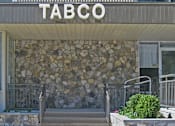 Thumbnail 1 of 10 - The front of a building with a stone wall and a sign at Tabco Towers Apartments, Towson, 21286