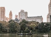 Thumbnail 16 of 17 - A boat with two people rowing on a lake in a park with trees and buildings in the background.