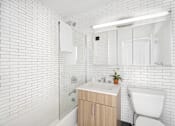 Thumbnail 25 of 56 - A white tiled bathroom with a wooden vanity and a white toilet.