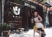 Thumbnail 51 of 56 - A woman in a yellow dress is walking past a coffee shop.