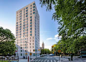 Thumbnail 1 of 9 - A tall building with many windows is surrounded by trees.