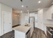Thumbnail 4 of 16 - Gallatin, TN, Apartments - The Gardens at Preston Park - Modern Kitchen with Vinyl Plank Flooring, Quartz Countertops, White Cabinetry, Stainless Steel Appliances, Tile Backsplash, Large Island, and Pendant Lights.
