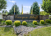 Thumbnail 47 of 47 - A sign that says Canyon Bluff in gold letters is surrounded by bushes and flags.