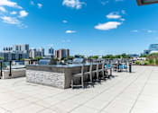 Thumbnail 22 of 24 - A rooftop patio with a bar and chairs overlooking a city skyline.