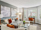 Thumbnail 9 of 34 - A living room with a grey couch, a glass table, and a painting on the wall.