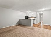 Thumbnail 7 of 25 - A kitchen with white cabinets and a black fridge is in the background of a living room with a beige carpet.