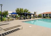 Thumbnail 31 of 46 - Mesa, AZ, Apartments with In-Unit Washer and Dryer - Mesa Station - Swimming Pool with Gazebo, Tanning Lounge Chairs, Tables, and Chairs Surrounded by Green Bushes.