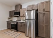 Thumbnail 1 of 18 - Modern kitchen at Blue Sky Landing featuring stainless steel appliances, wood-style cabinetry, efficient appliances, built-in microwave, dishwasher, and ample cabinet space in a brand-new College Park apartment.