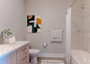 Thumbnail 11 of 22 - Modern bathroom at Creekline at Fairfield Apartments in Fairfield, CT featuring a soaking tub, marble-style tile surround, wood vanity, quartz countertop, and contemporary finishes.