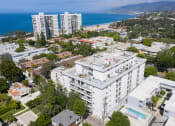 Thumbnail 22 of 29 - Apartments for Rent in Santa Monica, CA - San Vicente Tower - Aerial View of Property Exterior with White Walls, Surrounding Lush Green Neighborhood, Showcasing the Units and the Pool, with the Nearby Beach in View
