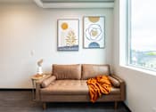 Thumbnail 31 of 54 - A tan couch with an orange throw is in front of a window with two framed pictures on the wall.
