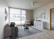 Thumbnail 2 of 20 - Two-Bedroom Apartments in South Seattle, WA - Greenhouse - Furnished Living Room with Carpeted Flooring, Area Rug, Grey Couch, Armchair, Coffee Table, Grey Cabinet, Ceiling Fan, House Plants, and Extra-Large Window.