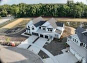 Thumbnail 8 of 14 - A white house with a black roof is surrounded by a driveway and a pond.