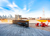 Thumbnail 12 of 29 - A wooden bench with black cushions is in the foreground of a rooftop with a city skyline in the background.