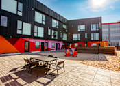 Thumbnail 23 of 29 - A wooden table and chairs are on a patio in front of a building with black and red walls.