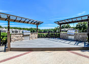 Thumbnail 8 of 17 - A wooden pergola with a stone wall and a building in the background.