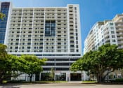 Thumbnail 18 of 19 - Two-Bedroom Apartments in Miami, FL - MB Station - Street View of Apartment Building with Trees, Grassy Lawns, Parking Garage Entrance, and Balconies.