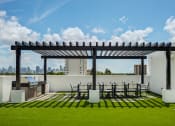 Thumbnail 6 of 19 - Apartments For Rent In Miami, FL - MB Station - Outdoor Grill Area With Pergola, Expansive Dining Table, Chairs, BBQ Grill, And A Grassy Courtyard