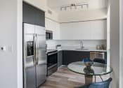 Thumbnail 1 of 19 - Studio Apartments in Miami, FL - MB Station - Modern Kitchen with Hardwood Flooring, Stainless Steel Appliances, White Upper Cabinetry, Grey Lower and Above-Fridge Cabinetry, Quartz Countertops, White Walls, Overhead Lighting, and Round Glass Top Dining Table with Two Chairs.