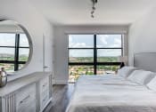 Thumbnail 2 of 19 - One-Bedroom Apartments in Miami, FL - MB Station - Furnished Bedroom with Hardwood Flooring, Large Bed, Grey Wooden Dresser, Framed Mounted Mirror, Nightstand with Lamp, Floor-to-Ceiling Window with City Views, and White Walls.