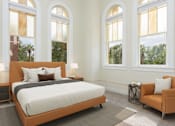 Thumbnail 5 of 39 - Affordable Apartments In San Antonio, TX, With In-Unit Washer And Dryer - The St. John - A Spacious Bedroom With Historic Arched Windows, A Tan Leather Platform Bed, And A Matching Accent Chair.