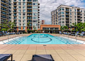 Thumbnail 10 of 25 - A large outdoor swimming pool with lounge chairs and apartment buildings in the background.