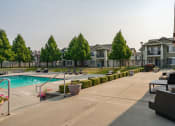 Thumbnail 12 of 26 - Apartments In Coeur d'Alene, ID, For Rent - Rockwood Lodge - Gated Resort-Style Pool With Expansive Sundeck, Lounge Seats, Manicured Hedgerow, Patio Area With Furniture, Trees, And Surrounding Apartment Buildings.
