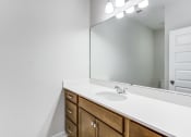Thumbnail 14 of 25 - Charleston, SC, Apartments - Sumner Village - Bathroom with Granite Vanity Countertop, Large Mirror, Bright Lights, and Wooden Cabinetry.