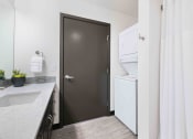 Thumbnail 21 of 50 - Greenwood, WA Apartments - The Morrow - Bathroom With In-Unit Washer And Dryer, Vinyl Plank Flooring, Granite Countertop, And Dark Brown Cabinets