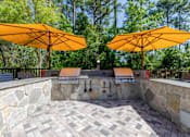 Thumbnail 13 of 32 - Outdoor grilling stations with umbrellas at The Sanctuary at 331 apartment complex in Santa Rosa Beach, Florida, surrounded by seating areas.