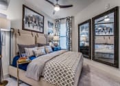 Thumbnail 7 of 18 - 	Dog Friendly Apartments in Grand Prairie, TX - Waters Edge at Mansfield - Bedroom With Custom Carpeting, Ceiling Fan, Window With 2" Blinds, Floor Lamp, Wall Art, Wall Mirrors, and Nightstand With La