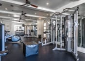 Thumbnail 8 of 40 - Studio Apartments in Rowlett, TX - Village of Rowlett - Fitness Center with Weights, Strength Training Machines, Ceiling Fans, Large Mirrors, and Windows.