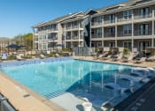 Thumbnail 19 of 40 - Two-Bedroom Apartments in Rowlett, TX - Village of Rowlett - Gated Resort-Style Swimming Pool with Submerged and Poolside Lounge Seats, Umbrellas, and Building Exterior in Background.