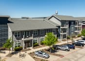 Thumbnail 29 of 40 - Apartments for Rent Rowlett, TX - Village of Rowlett - Aerial View of Property Exterior with Grey and Blue Walls, Showcasing Nearby Trees and Parking Spaces.
