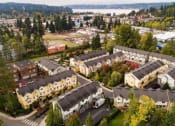 Thumbnail 7 of 14 - Apartments for Rent in Kenmore, WA - Northshore Townhomes - Aerial View of the Property Featuring Tan and Yellow Buildings with Grey Roofing, Surrounded by Lush Nature and Landscaping, with Nearby Lake in the Background.