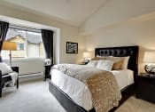 Thumbnail 5 of 14 - Three Bedroom Apartments in Kenmore WA - Northshore Townhomes - Furnished Bedroom with a Large Bed, Carpeted Floor, Black Nightstands with Lamps, an Armchair, Window, and Standing Lamp